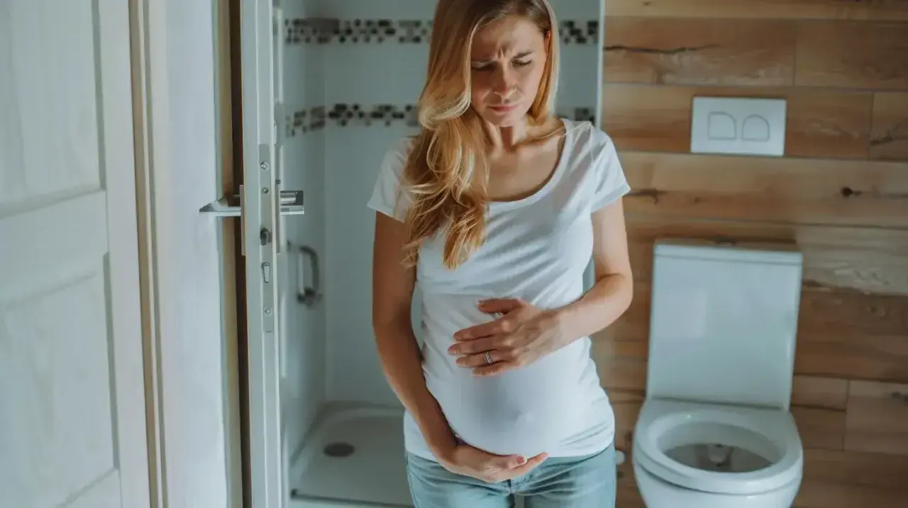 Kobieta w ciąży trzyma się za brzuch, cierpiąc z powodu bólu podbrzusza. W tle łazienka z toaletą.