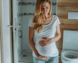 Kobieta w ciąży trzyma się za brzuch, cierpiąc z powodu bólu podbrzusza. W tle łazienka z toaletą.