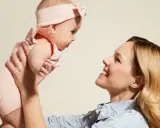 Mama podnosi dziecko w różowym body i opasce. Idealny przykład, jak ubierać noworodka latem – lekko i wygodnie.