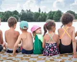 Szczęśliwe wakacje nad jeziorem z dziećmi. Grupa dzieci siedzi na kocu, ciesząc się słońcem i widokiem na wodę.