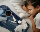 Chory chłopiec śpi, obok kubek z naparem, termometr, maska i chusteczki. To objawy grypy A.