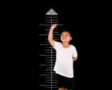 Uśmiechnięty chłopiec mierzy swój wzrost na tle czarnej tablicy z zaznaczoną miarką. Ma 135 cm wzrostu i siedzi bez fotelika.