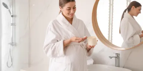 Kobieta w ciąży, w białym szlafroku, aplikuje krem. Bezpieczne kosmetyki, jak te bez kwasu salicylowego w ciąży, to podstawa.