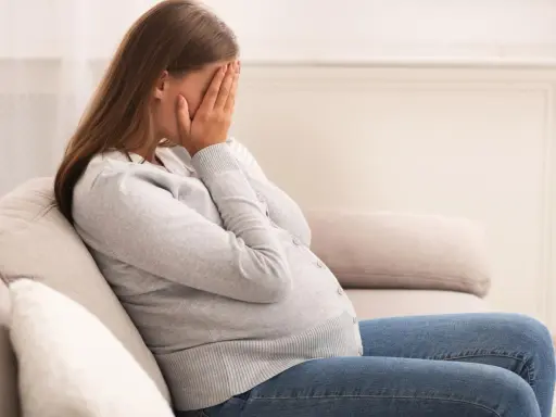 Kobieta w ciąży siedzi na kanapie, zakrywając twarz dłońmi. Jej postawa wyraża głęboki smutek, być może związany z diagnozą wady letalnej.