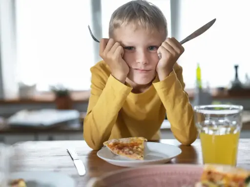 Chłopiec z widelcem i nożem w dłoniach, z miną mówiącą "dziecko nie chce jeść". Przed nim kawałek pizzy i szklanka soku.