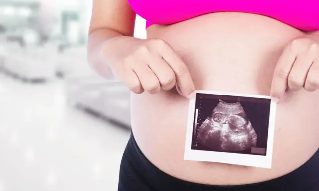 Ciężarna kobieta trzyma zdjęcie USG płodu na tle rozmazanego wnętrza.