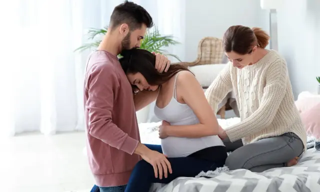 Mężczyzna obejmuje ciężarną kobietę, która opiera głowę na jego ramieniu. Obok siedzi doula, która delikatnie dotyka jej brzucha, oferując wsparcie.