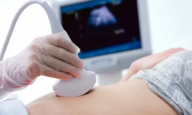 Ręka w rękawiczce trzyma sondę USG, która bada brzuch. Na ekranie widać obraz z badania, być może to test pappa.