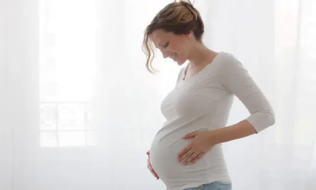 Uśmiechnięta ciężarna kobieta na urlopie macierzyńskim delikatnie dotyka brzucha.
