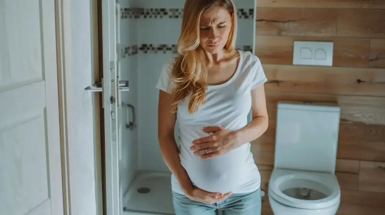 Kobieta w ciąży trzyma się za brzuch, cierpiąc z powodu bólu podbrzusza. W tle łazienka z toaletą.