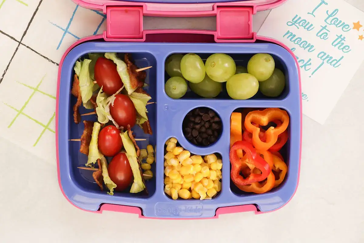 Kolorowy lunchbox z przekąskami dla dzieci: szaszłyki z pomidorków i boczku, winogrona, fasolka, kukurydza i papryka.