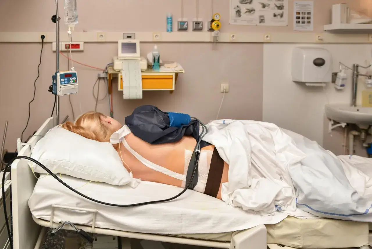 Pacjentka w łóżku szpitalnym, podłączona do aparatury medycznej. Widoczny cewnik i opatrunki na plecach, sugerujące podanie znieczulenia epiduralnego.