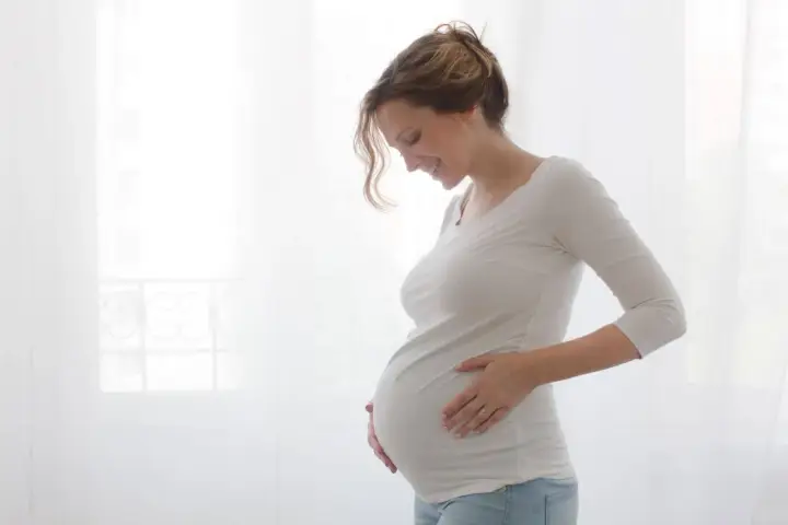 Uśmiechnięta ciężarna kobieta na urlopie macierzyńskim delikatnie dotyka brzucha.